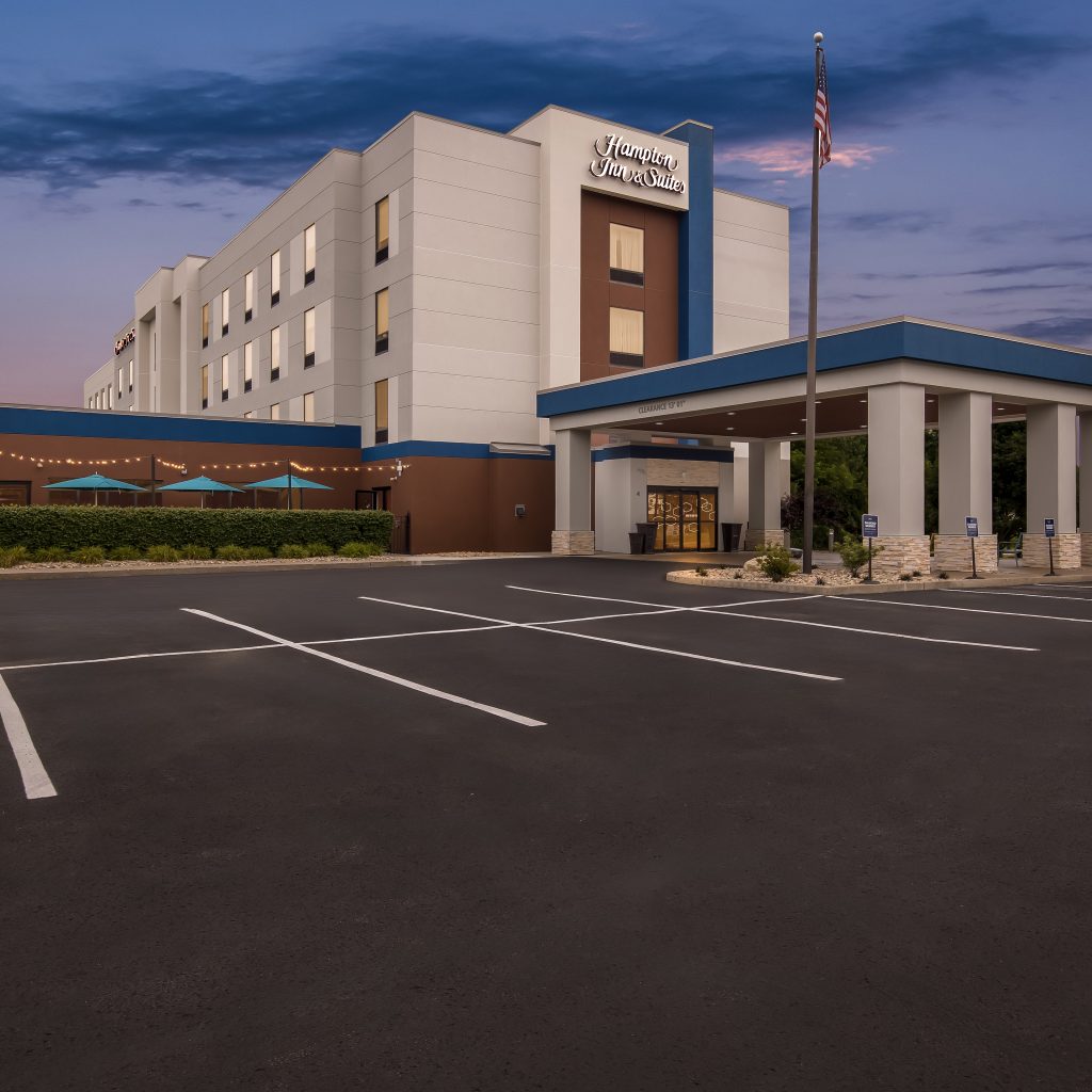 Front view of a Hampton Inn & Suites hotel with a covered entrance, blue trim, and a paved parking lot at dusk.