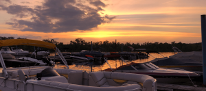 Boats docked at a marina at sunset with an orange sky and calm water reflection.
