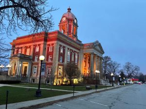 Red brick courthouse with a domed clock tower, illuminated at dusk, featuring neoclassical columns and a lamps-lit street.