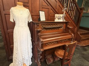 Wooden carved piano with a white lace wedding dress on a mannequin beside it, in a museum display with photos and a staircase in the background.