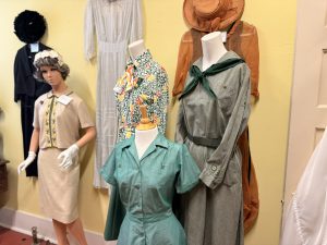 Mannequins wearing vintage dresses and hats on display against a pale yellow wall in a shop window.