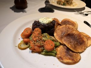 Plate of beef steak topped with herb butter, beside roasted carrot coins and potato pancakes on a white tablecloth plate with a side of vegetables.