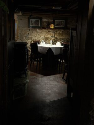 Dimly lit dining nook with stone wall, round table set for two with folded napkins and glasses, framed pictures above.