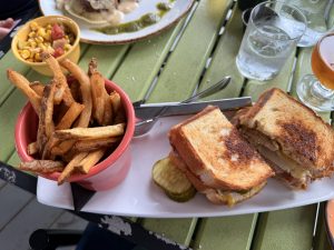 Meal spread on a green table: fries in a pink cup, corn salad, a toasted cheese sandwich, pickles, and drinks.