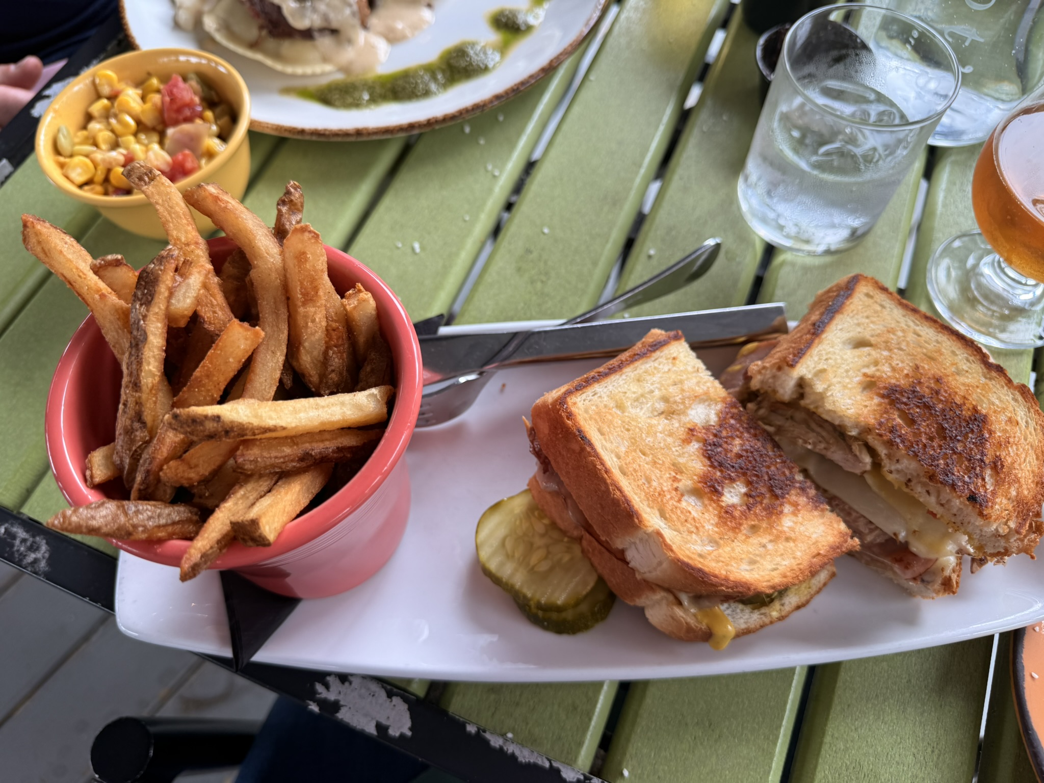 Meal spread on a green table: fries in a pink cup, corn salad, a toasted cheese sandwich, pickles, and drinks.