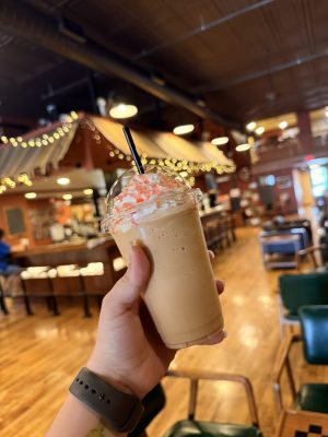 Person holding a whipped-cream frappé with pink drizzle in a clear cup inside a cozy cafe.