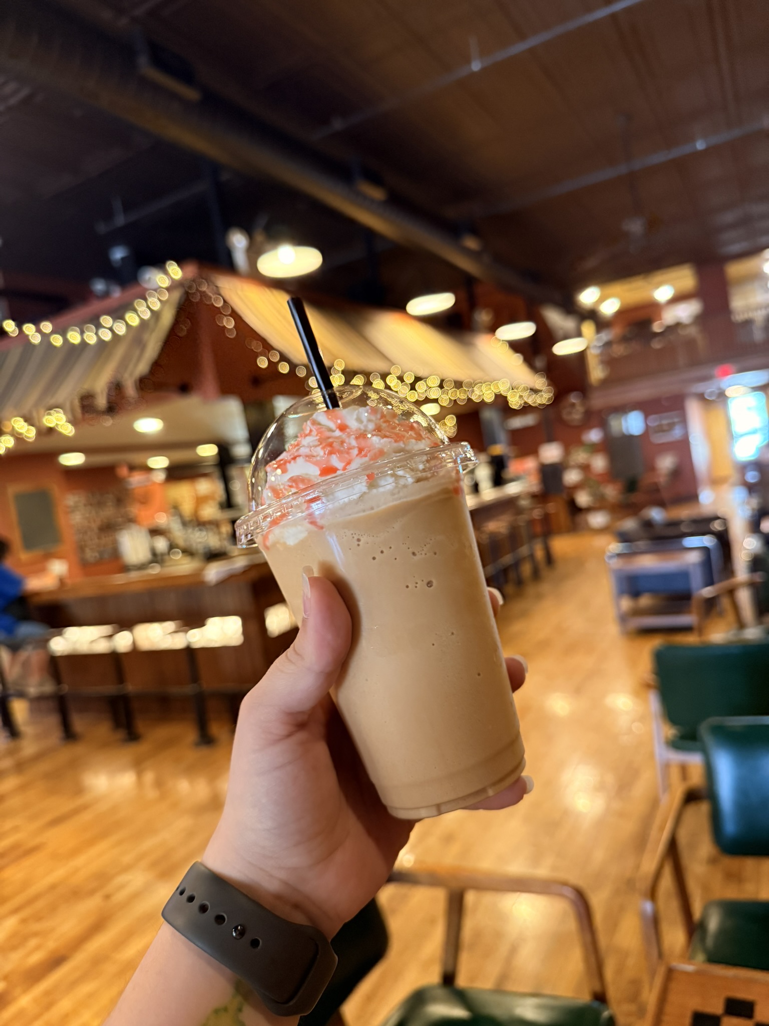 Person holding a whipped-cream frappé with pink drizzle in a clear cup inside a cozy cafe.