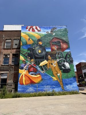 Large mural on a brick building showing a colorful train and rural landscape under a blue sky, with people and animals in the scene.