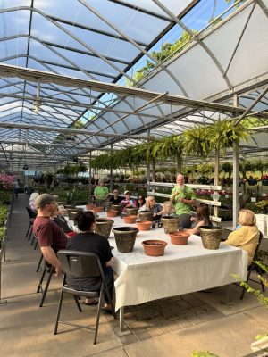 Group of people seated around a long table in a glass greenhouse, with many clay pots in front, a man in a green shirt speaks at the head of the table.