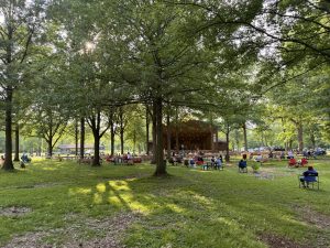 Outdoor park concert under large trees, audience seated on benches facing a stage.