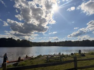 Calm lake at a park with kayaks on the shore, a wooden fence, and people near the water under a sunny sky with clouds.