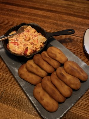 Cast-iron skillet with creamy potato dish and red peppers, spoon resting inside, on a wooden table, next to a tray of golden dinner rolls.