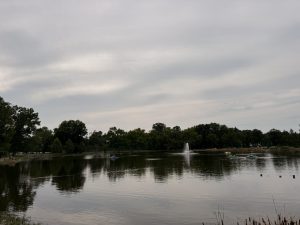 A lake with a central fountain surrounded by trees and small boats on the water under a cloudy sky.