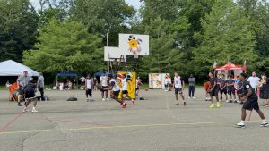 Outdoor basketball game with players in white and dark jerseys, officials in stripes, and tents along the sideline under green trees.