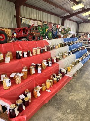 Jars of homemade preserves lined on red, white, and blue tables with vintage toy tractors and quilts on display in a barn-like market stall interior.