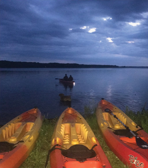 Evening scene at a lake: three colorful kayaks on the shore with two people paddling in a distant boat under a cloudy blue sky.