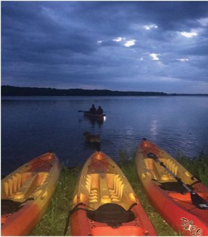 Three orange kayaks on the grassy shore face a calm lake as two people paddle a small boat under a blue, cloudy dusk sky.