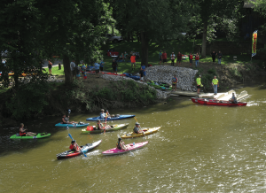 Colorful kayaks paddle down a brown river as spectators on the rocky riverbank watch nearby start/finish banners in the background.