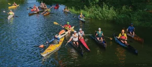 A large group of kayakers in colorful boats paddling together on a sunlit river.