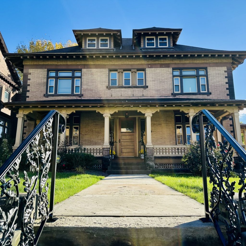 Brick two-story house with a front porch and decorative black metal railings along the walkway to the entrance on a sunny day