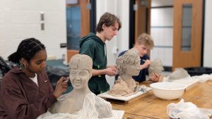 Students in a sculpture class shaping clay busts at a long table, focused on their work with bowls of clay nearby.