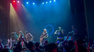Band performing on a lit stage with colorful spotlights and a cheering crowd.