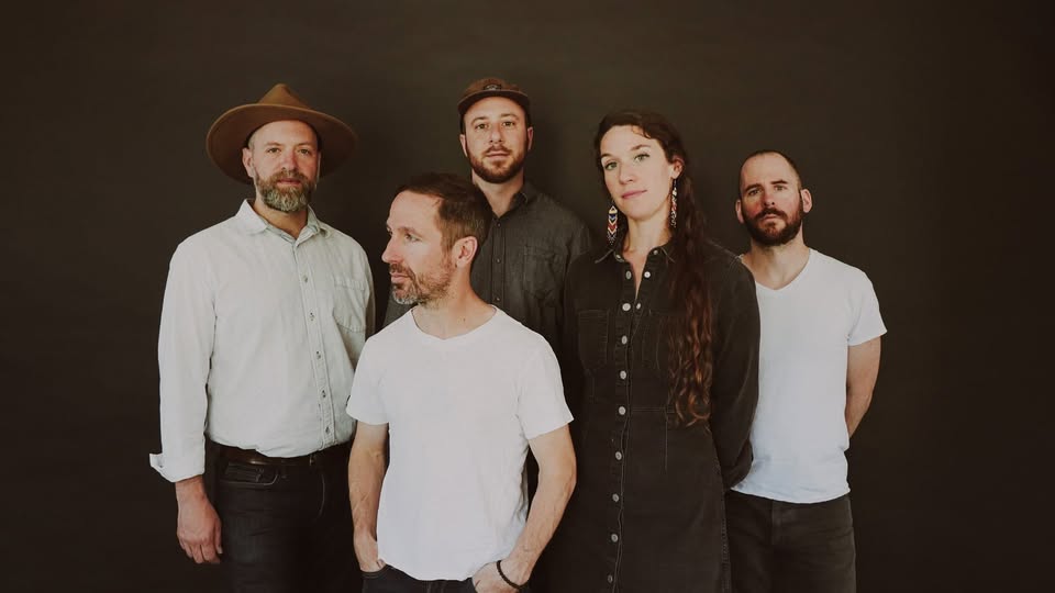 Five adults posing together in a studio against a dark backdrop; two men wear white tops, one man in a hat, a woman in a dark dress, and another man in a white t-shirt.