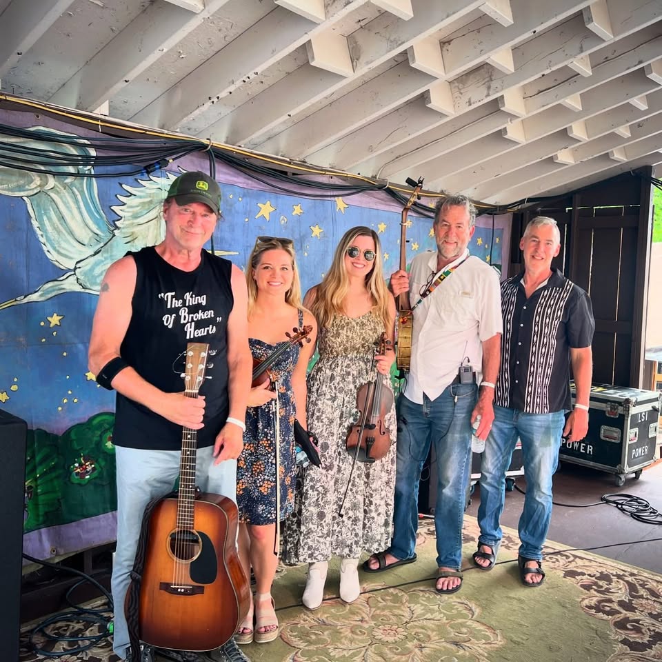 Five musicians posing on a stage: guitarist, two violinists, and two others with instruments, in front of a mural backdrop with a blue sky and stars.