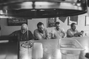 Four men sit at a bar counter in a grayscale cafe, some looking at drinks, a bottle visible on the right edge.