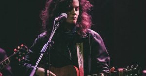 Musician with long hair and a leather jacket playing an acoustic guitar on a dimly lit stage, singing into a microphone.