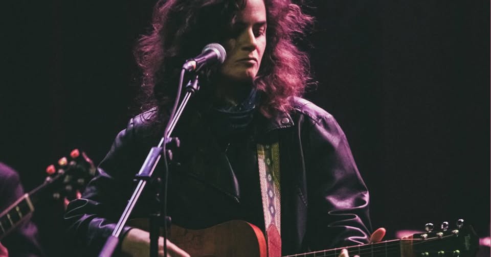 Musician with long hair and a leather jacket playing an acoustic guitar on a dimly lit stage, singing into a microphone.