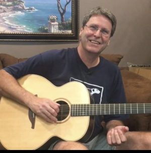 Smiling man sits on a brown couch, strumming a light-colored acoustic guitar indoors with a landscape painting in the background.