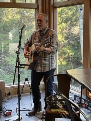 Male musician playing an acoustic guitar at a microphone in a sunlit room with large windows and trees outside.