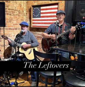 Two men playing acoustic guitars and singing into microphones on a brick-wall stage, with an American flag backdrop and a banner reading 'The Leftovers'.