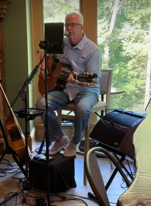 Man with glasses sings into a microphone while playing an acoustic guitar in a sunlit home studioed room with a window view behind him.