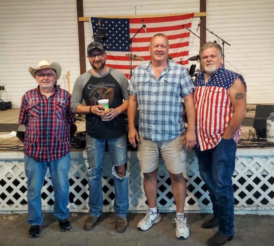 Four men standing side by side on a stage with a U.S. flag backdrop; one holds a cup of beverage.