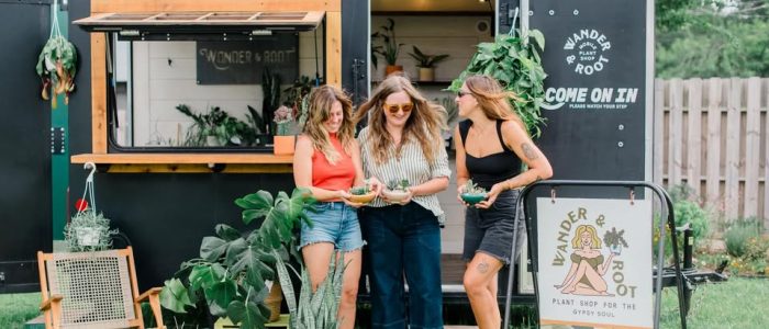 Three women stand in front of a black mobile plant shop wagon, holding small potted plants and smiling outdoors.