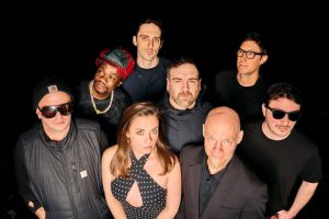 Eight-person group portrait of a band in dark clothing posed against a black backdrop, with a woman in a polka-dot dress center-front.