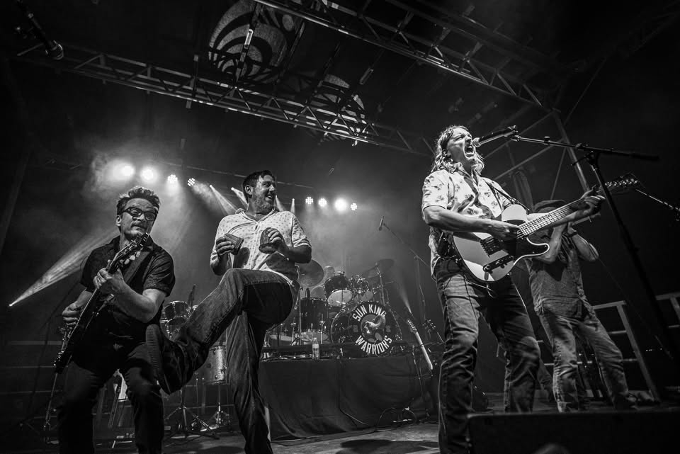 Black-and-white photo of a four-member rock band performing on a lit stage, with guitarists on the sides and drums in the back under bright lights, conveying a high-energy live show.