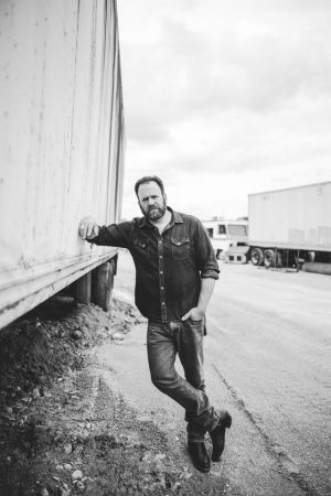 Bearded man in a denim shirt and jeans leaning against a large truck trailer in a dusty yard (black-and-white).