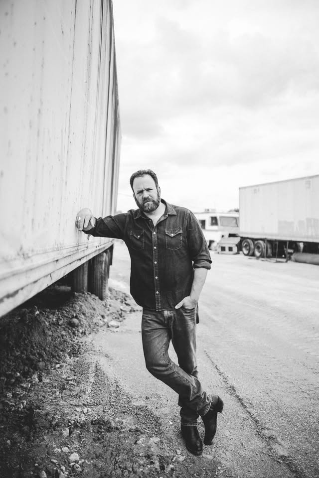 Bearded man in a denim shirt and jeans leaning against a large truck trailer in a dusty yard (black-and-white).