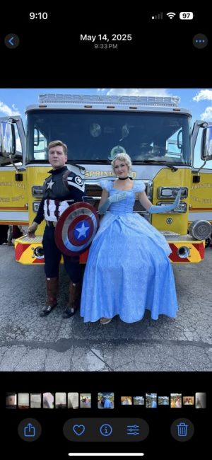 Two costumed figures, Captain America and Cinderella, pose in front of a bright yellow fire truck on a sunny day.