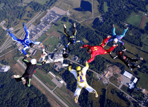 Group of skydivers in colorful suits forming a circular formation in freefall above a rural landscape with fields and buildings below