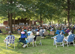 Outdoor park concert with a crowd seated in lawn chairs facing a small stage under trees.] ,
