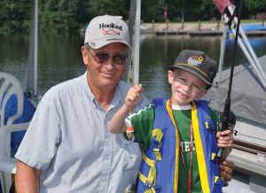 Grandfather and grandson posing on a boat by the water after a fishing trip, the boy proudly holding a small fish on the line while the man smiles nearby.
