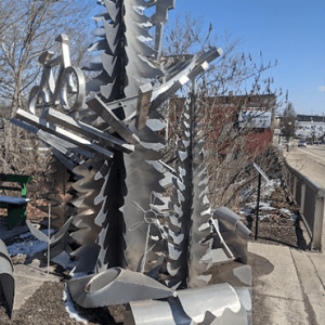 Abstract outdoor sculpture built from stacked saw blades and metal beams against a blue sky
