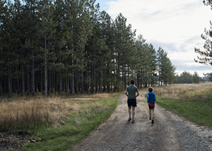 Two people jogging on a gravel trail through a forested area, an adult and a child side by side.