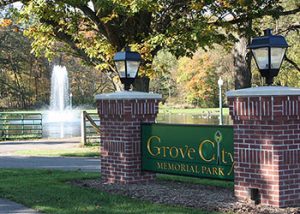 Brick entrance pillars with a green Grove City Memorial Park sign, lampposts, and a fountain in a tree-filled park.