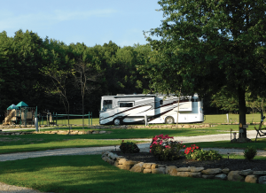 A white motorhome parked on a campground road with a playground and trees nearby in a sunny park.