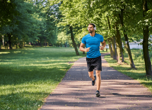 Man in a blue T-shirt and black shorts jogs along a sunlit, tree-lined park path wearing headphones.
